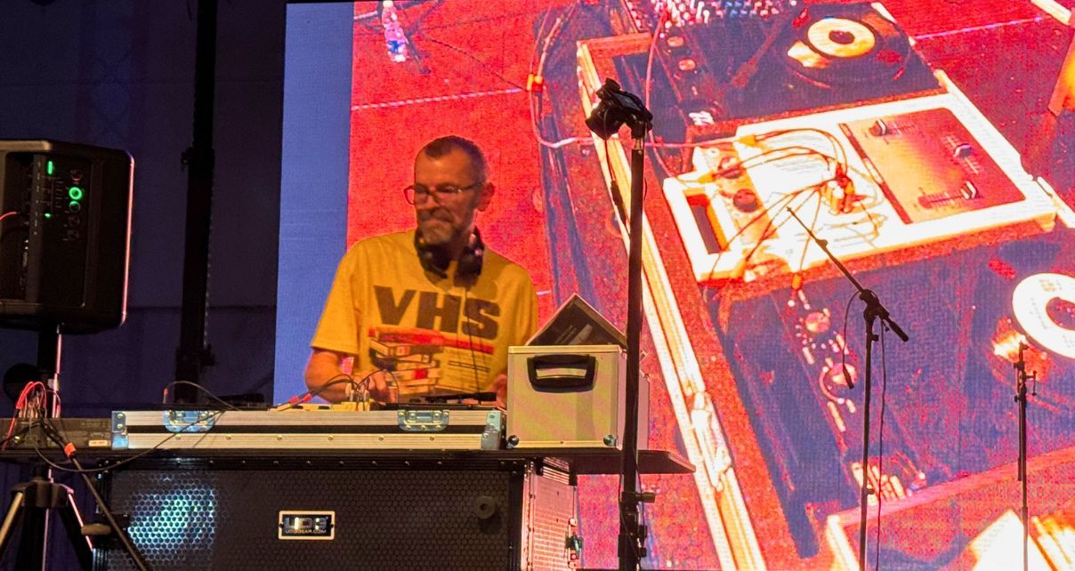 A white man with a beard, shaved hair, and glasses stands on a stage behind a DJ booth. He's wearing black headphones and a white shirt that says "VHS" in big letters. Behind him you can see retro DJ gear displayed on a big screen.