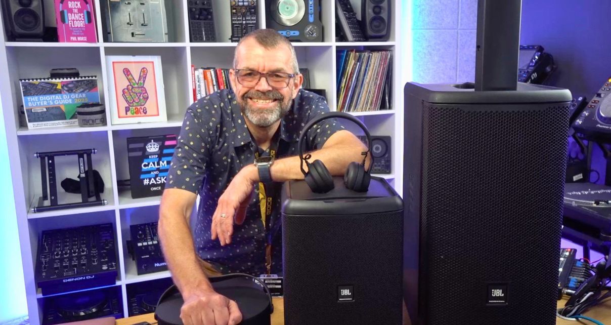 A white person in their 50s with a greying beard, close-shaved head, and glasses leans forward and smiles at the camera from behind a wooden desk. In the background is a large white bookshelf holding various books, DJ gear, and artwork. In the foreground, there are several black speakers and a pair of headphones sitting on the desk.