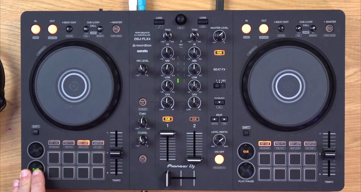 Top-down view of a black AlphaTheta DDJ-FLX2 DJ controller sitting on a wooden desk. In the lower left-hand corner you can see a white person pressing the Start button with two fingers.