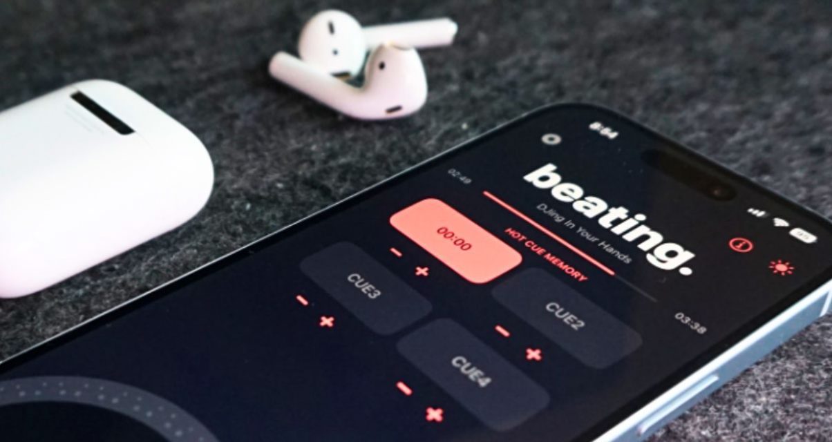 An iPhone sits next to a pair of AirPods on a grey surface. The phone is displaying a DJ app called "beating.", which has a black, dark grey, and light red colour scheme.