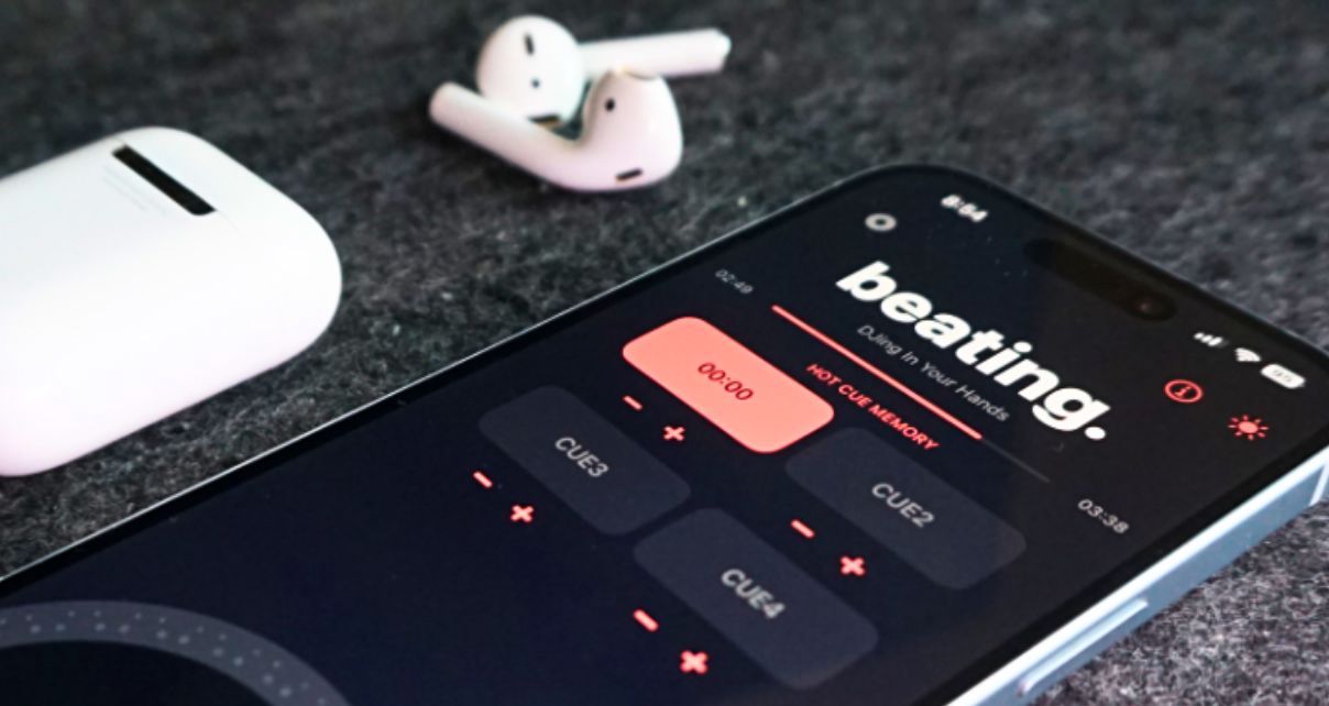 An iPhone sits next to a pair of AirPods on a grey surface. The phone is displaying a DJ app called "beating.", which has a black, dark grey, and light red colour scheme.