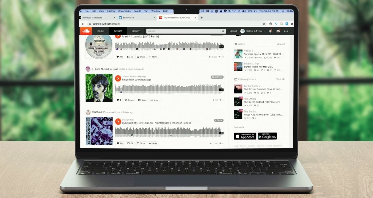 A silver laptop sits on a light wooden table against a lush green background. The screen displays a SoundCloud stream page featuring various audio tracks, play buttons, and waveform visualisations.