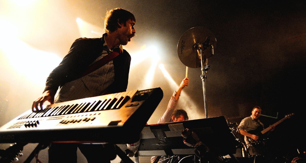 A lively band performs on stage, with musicians playing keyboard, drums, and guitar. Everything is illuminated by dramatic stage lighting.