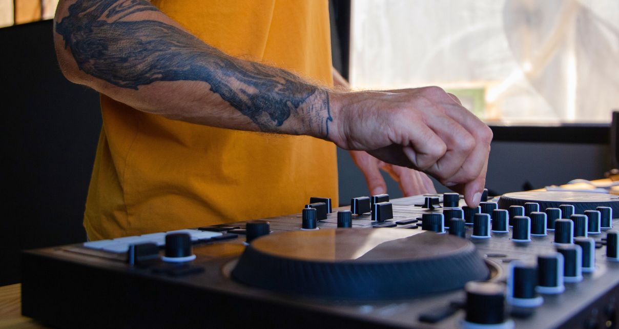A tattooed DJ in a yellow shirt adjusts knobs on a mixer, focusing intently on mixing tracks in a bright studio setting.