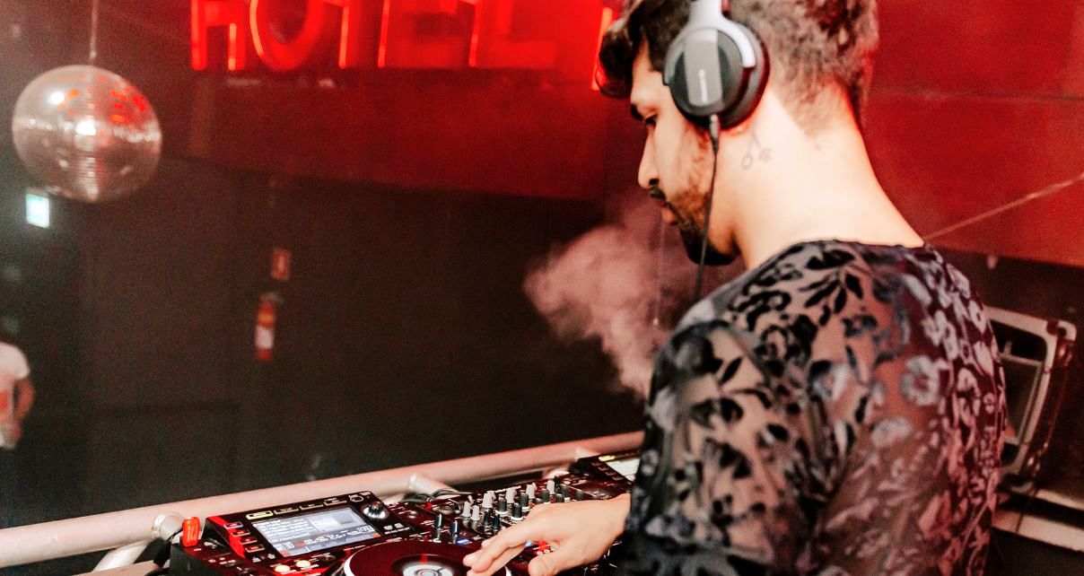 A youthful DJ stands behind a pro Pioneer DJ setup in a nearly empty nightclub. The DJ booth is surrounded by vibrant red lighting, and there's a disco ball off in the distance.
