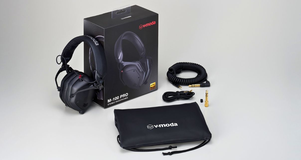 Black V-Moda M-100 Pro headphones displayed with their packaging. Sitting next to them on a light grey table are the detachable coiled and straight cables, a gold adapter, and a black carrying pouch.
