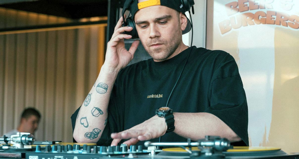 A white, tattooed DJ in a backwards baseball hat and black t-shirt mixes music on turntables in a casual setting. In the background you can see a person sitting down and a sign that says "Beer & Burgers".