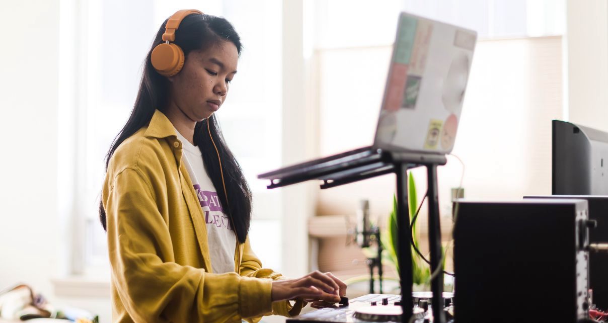 A person with long dark hair, bright orange headphones, and a yellow collared shirt stands behind a DJ controller and laptop in a well-lit room.