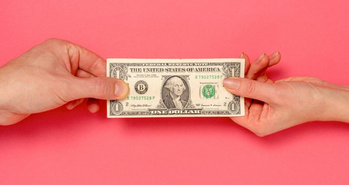 Two hands exchanging a crisp one-dollar bill against a vibrant pink background, highlighting a moment of transaction.