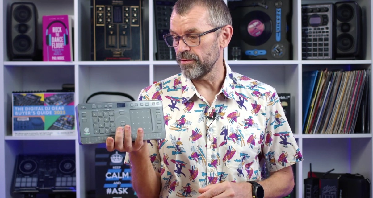 Phil Morse stands in the Digital DJ Tips studio against a bookshelf filled with DJ gear, music books, records, and more. 

Phil is a white person in his mid-50s with a salt-and-pepper beard, dark-rimmed glasses, close-shaved hair and is wearing a colourful patterned button-up t-shirt. 

He is flooking down and holding a sleek grey Chordcat groovebox unit. 