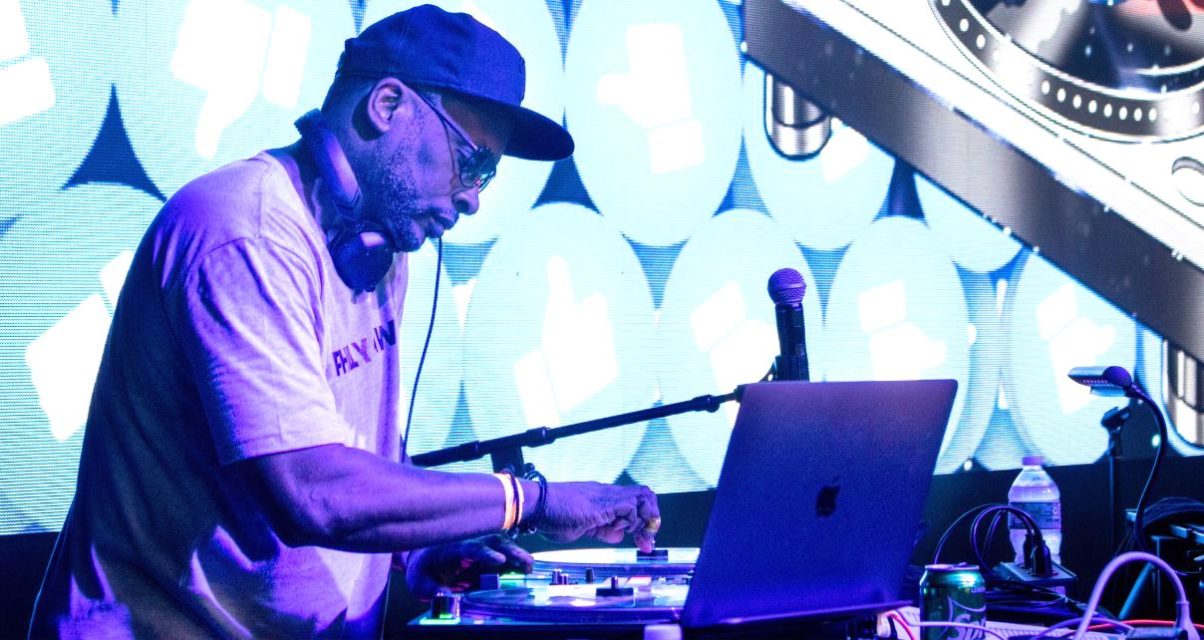DJ Jazzy Jeff performs on stage, mixing tracks with turntables and a laptop, illuminated by vibrant blue digital graphics in the background.