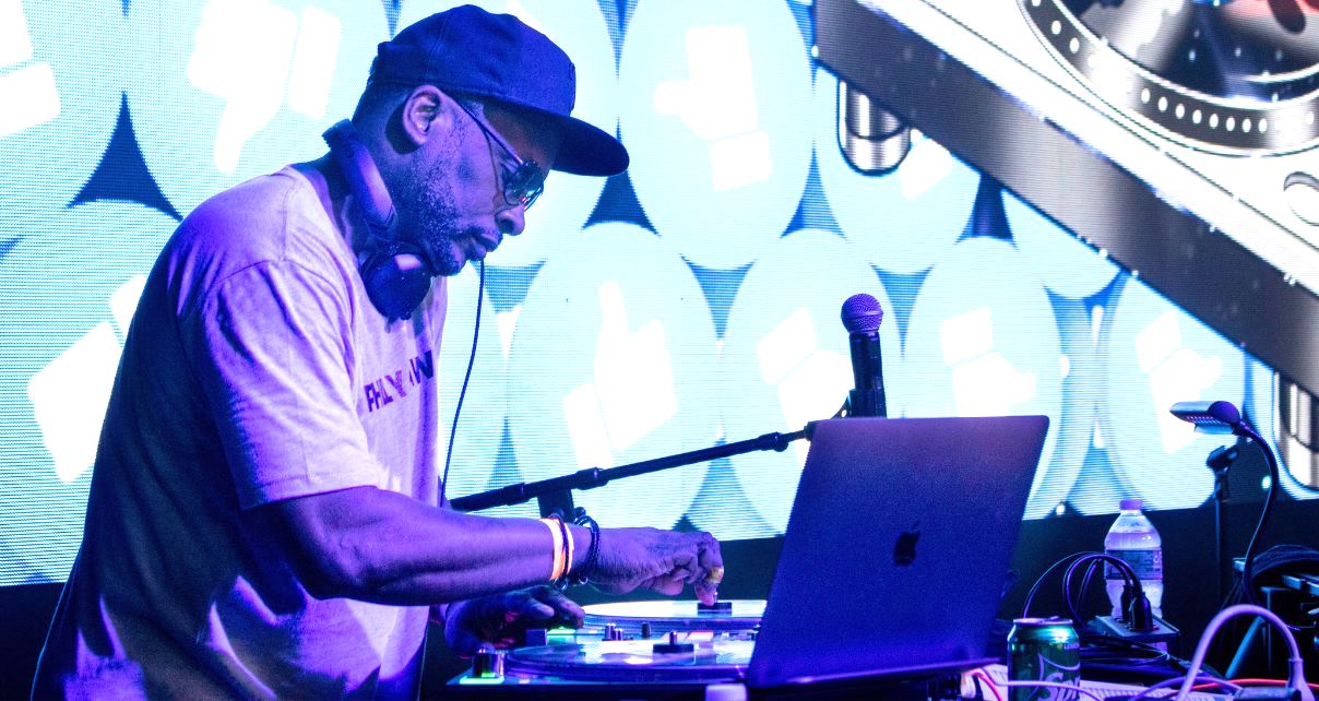 DJ Jazzy Jeff performs on stage, mixing tracks with turntables and a laptop, illuminated by vibrant blue digital graphics in the background.