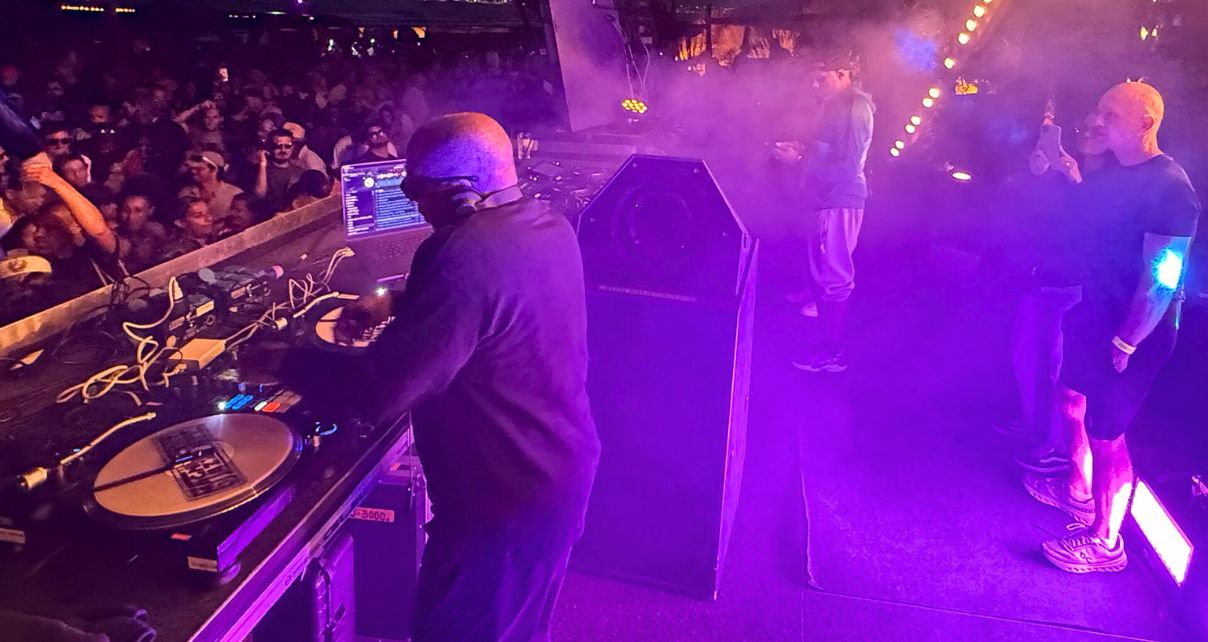 DJ Jazzy Jeff performs on stage at a beachside gig. Purple lighting illuminates the stage while an enthusiastic crowd enjoys the music in the background.