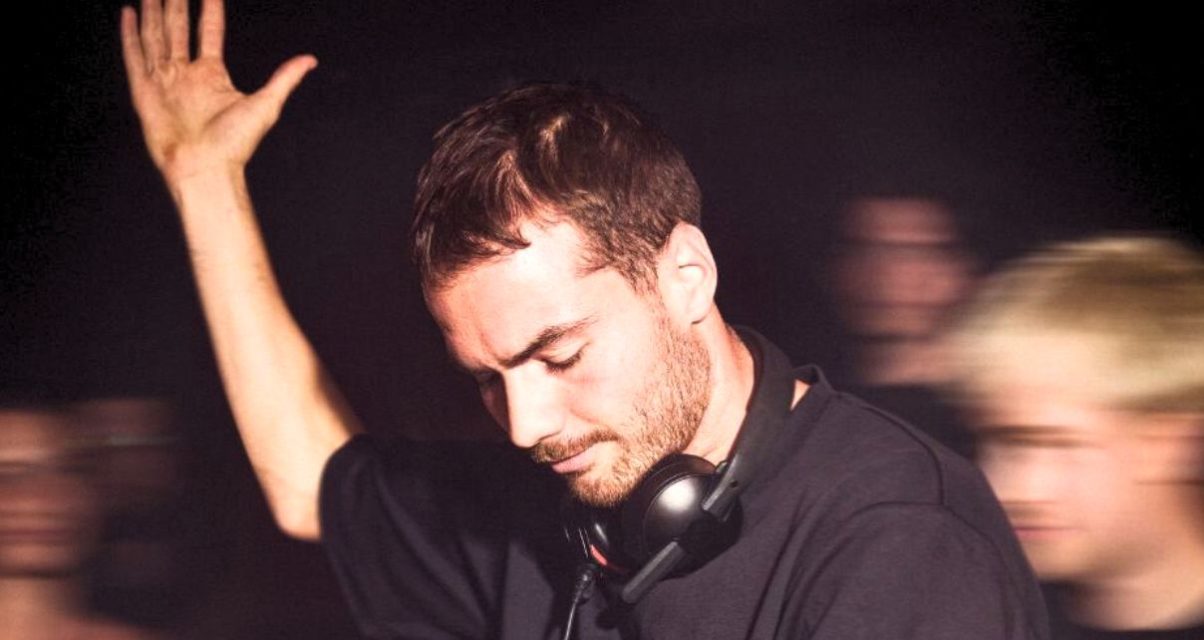 A white male-presenting DJ with short dark hair and a beard raises his hand while performing in a lively, dimly lit venue, surrounded by an energetic crowd. He is wearing a black t-shirt with a pair of black Røde NTH-50 headphones around his neck.