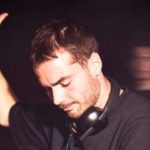 A white male-presenting DJ with short dark hair and a beard raises his hand while performing in a lively, dimly lit venue, surrounded by an energetic crowd. He is wearing a black t-shirt with a pair of black Røde NTH-50 headphones around his neck.