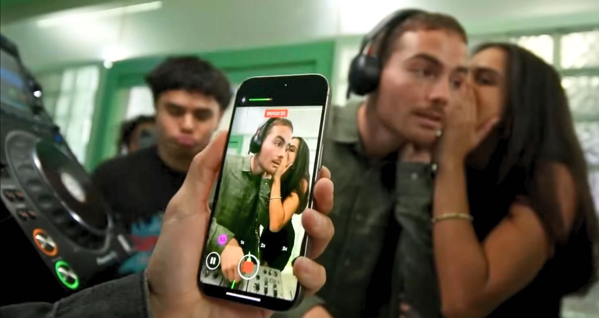 A hand holds a phone recording a lively moment in the DJ booth. Someone whispers into the DJ's ear, and several others enjoy the music in a vibrant, green-lit space.