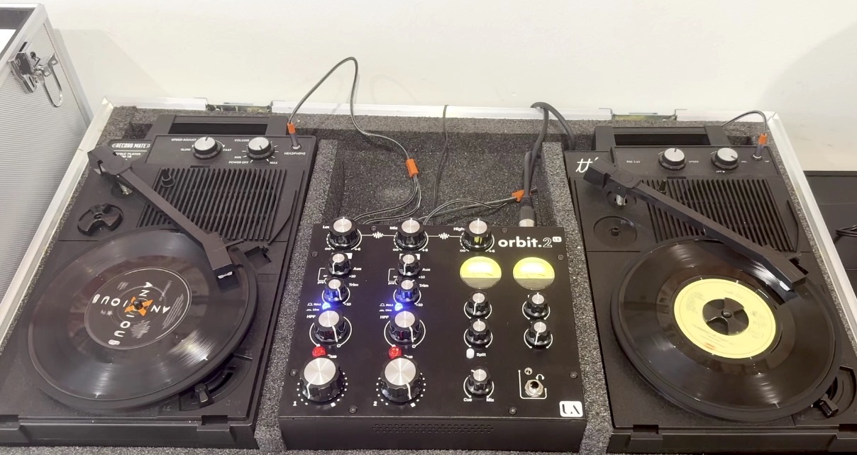 Top-down view of an all-analogue DJ set-up inside a silver case. The black Union Audio Orbit.2 LE mixer features various rotary knob controls, two yellow VU metres, and blue and red indicator lights. It sits between two portable black Record Mate turntables.