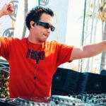 Laidback Luke is wearing a red t-shirt and sunglasses while DJing on an outdoor stage. He's raising both arms in excitement against a backdrop of palm trees and vibrant graphics.