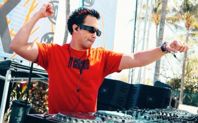 Laidback Luke is wearing a red t-shirt and sunglasses while DJing on an outdoor stage. He's raising both arms in excitement against a backdrop of palm trees and vibrant graphics.