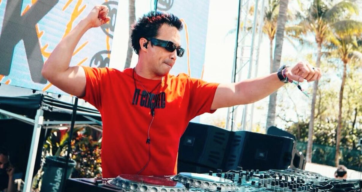 Laidback Luke is wearing a red t-shirt and sunglasses while DJing on an outdoor stage. He's raising both arms in excitement against a backdrop of palm trees and vibrant graphics.
