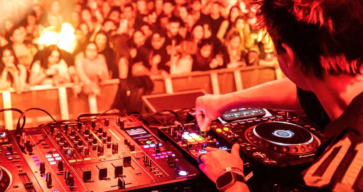 Laidback Luke in the DJ booth performing to a crowd of people at a festival. He's looking at the crowd and tweaking the controls a Reloop modular controller.