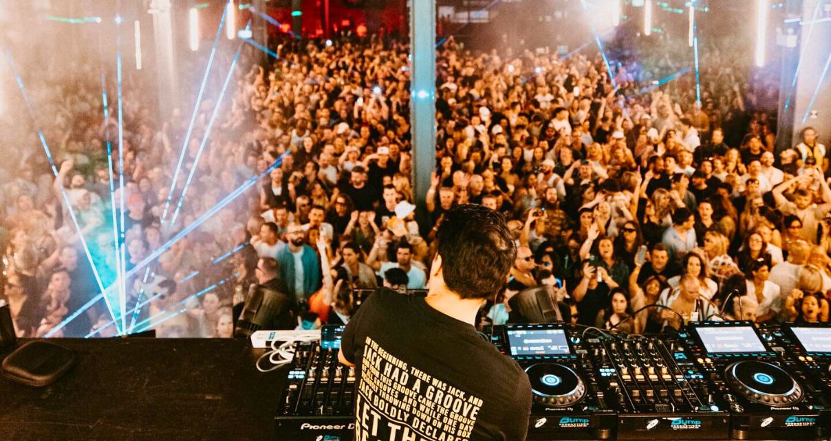 Laidback Luke stands behind a pro DJ booth in a well-lit warehouse, facing a huge crowd illuminated by colourful lasers and lights.
