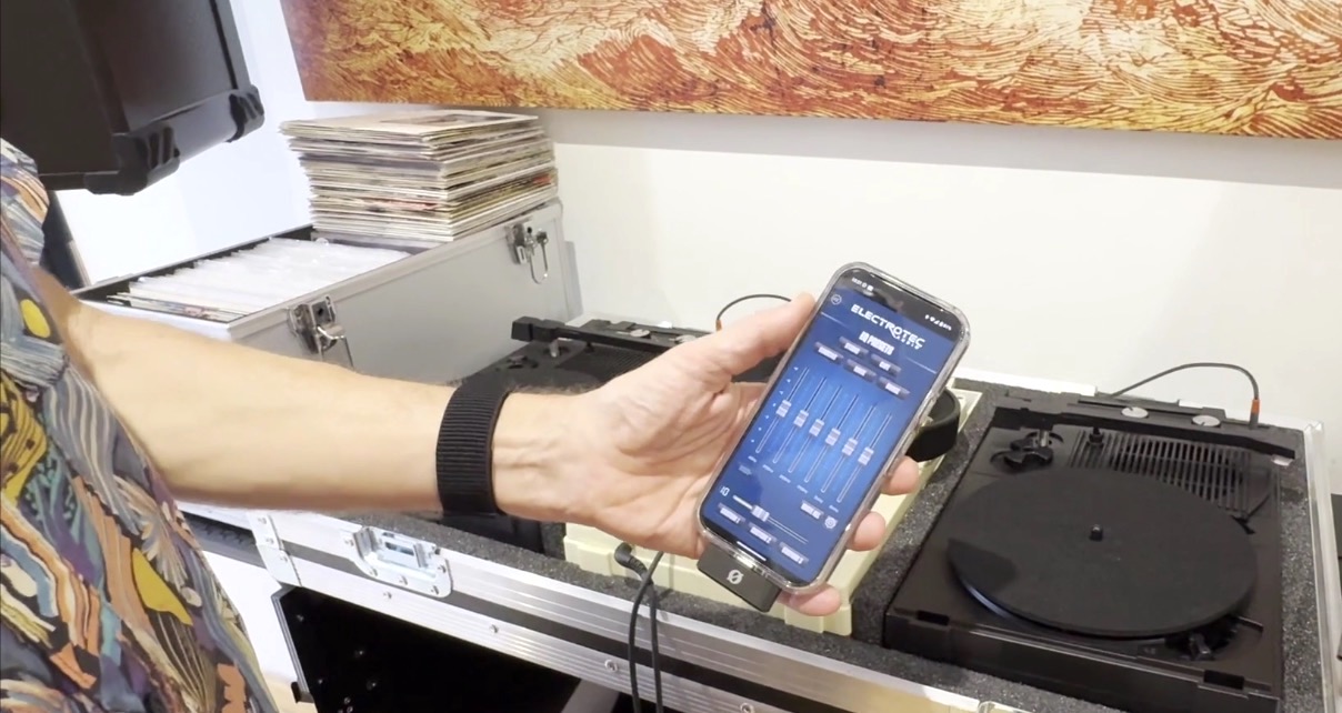 A white person wearing a colourful patterned shirt and black wristwatch holds a smartphone displaying the EQ settings on an audio mixing app, next to a turntable and vinyl records.