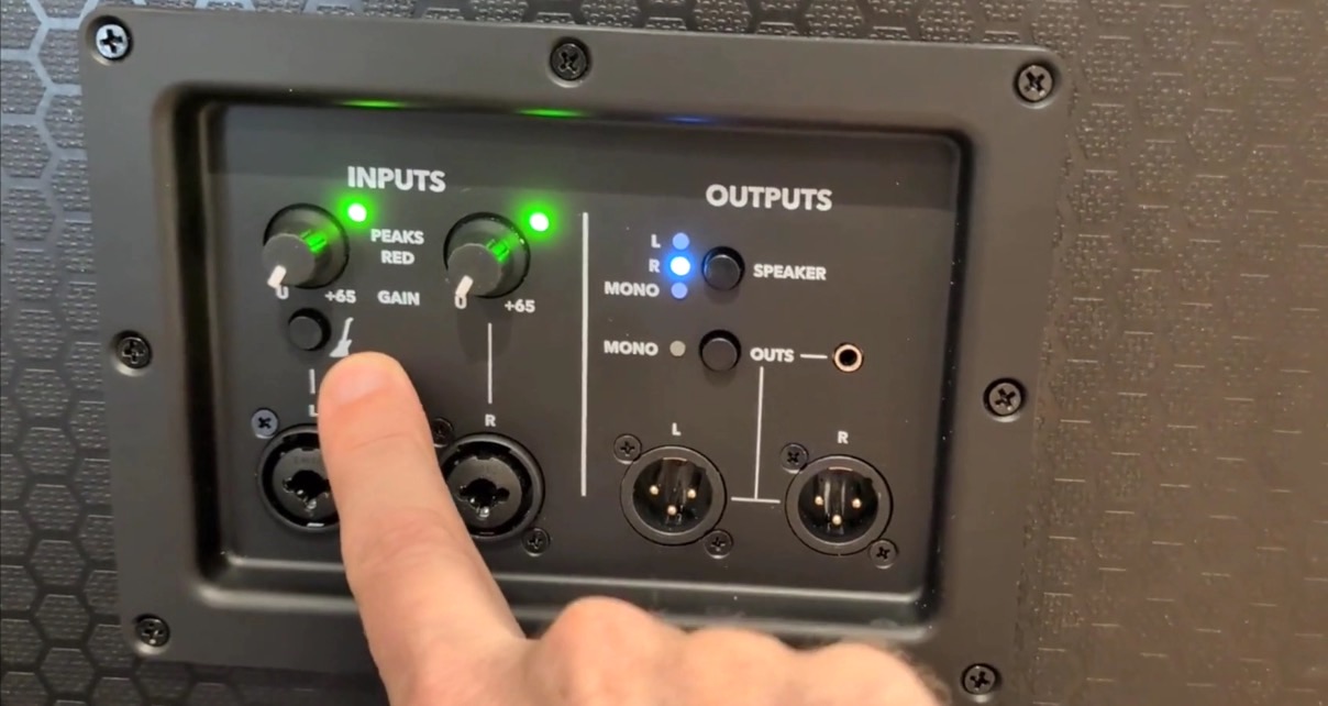 A hand points to the input controls on a rear speaker panel. It's displaying gain knobs and input and output connections, with green and blue LED indicators lit. 