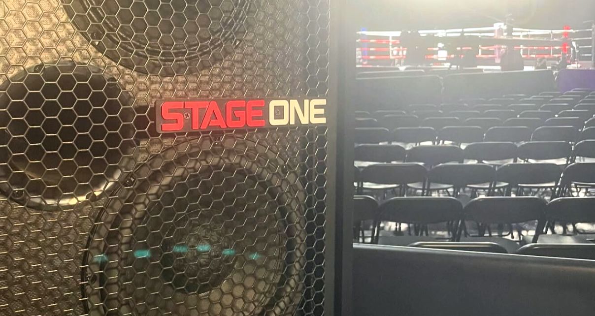 Close-up of a "Stage One" speaker with a boxing ring and empty chairs in the background, set for an event in a dimly lit venue. 