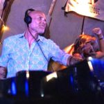 A DJ in a floral shirt performs in a pro DJ booth at a lively venue, with lights twinkling and a dancer raising her arm in enjoyment.
