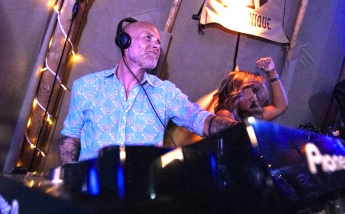 A DJ in a floral shirt performs in a pro DJ booth at a lively venue, with lights twinkling and a dancer raising her arm in enjoyment.