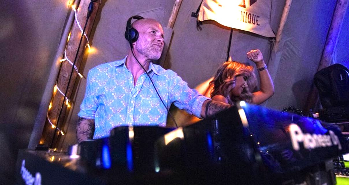 A DJ in a floral shirt performs in a pro DJ booth at a lively venue, with lights twinkling and a dancer raising her arm in enjoyment.