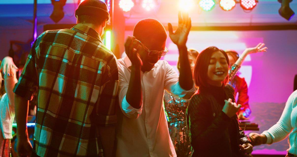A vibrant dance party scene featuring a diverse group of people dancing and smiling under colourful lights. 