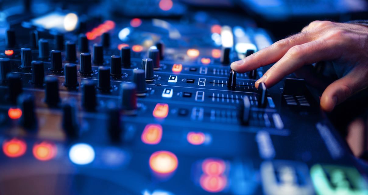 A close-up of a DJ's hand adjusting the volume levels on a colourfully lit DJ mixer. 