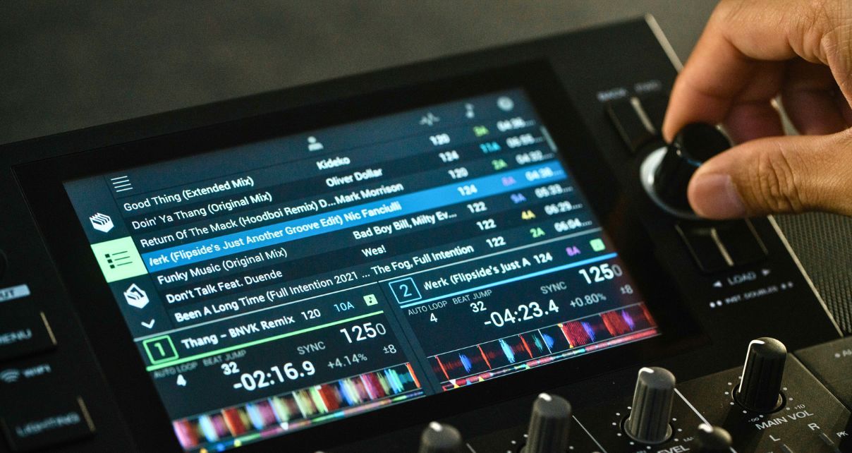 Close-up of a DJ controller’s screen showing various music tracks and settings, with a hand on the right adjusting the load knob.