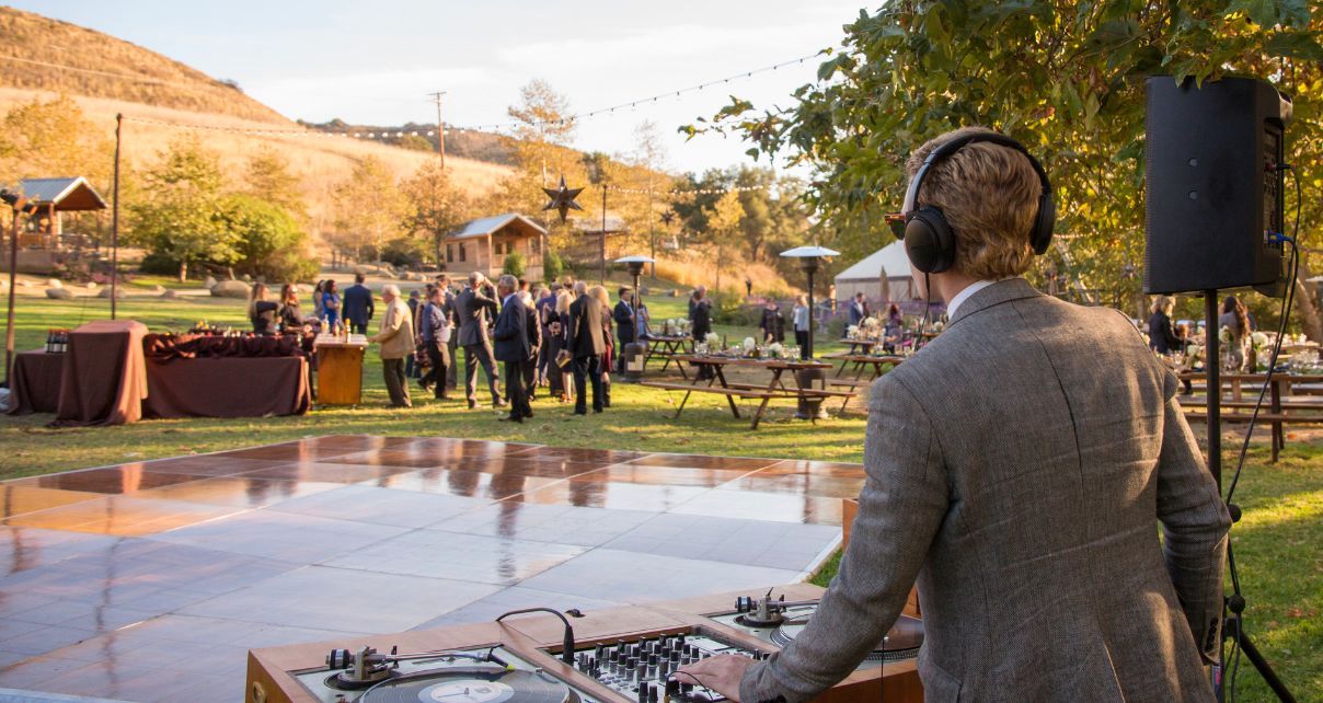 A DJ in a tan suit mixing music at an outdoor event with guests mingling in a scenic garden setting. 