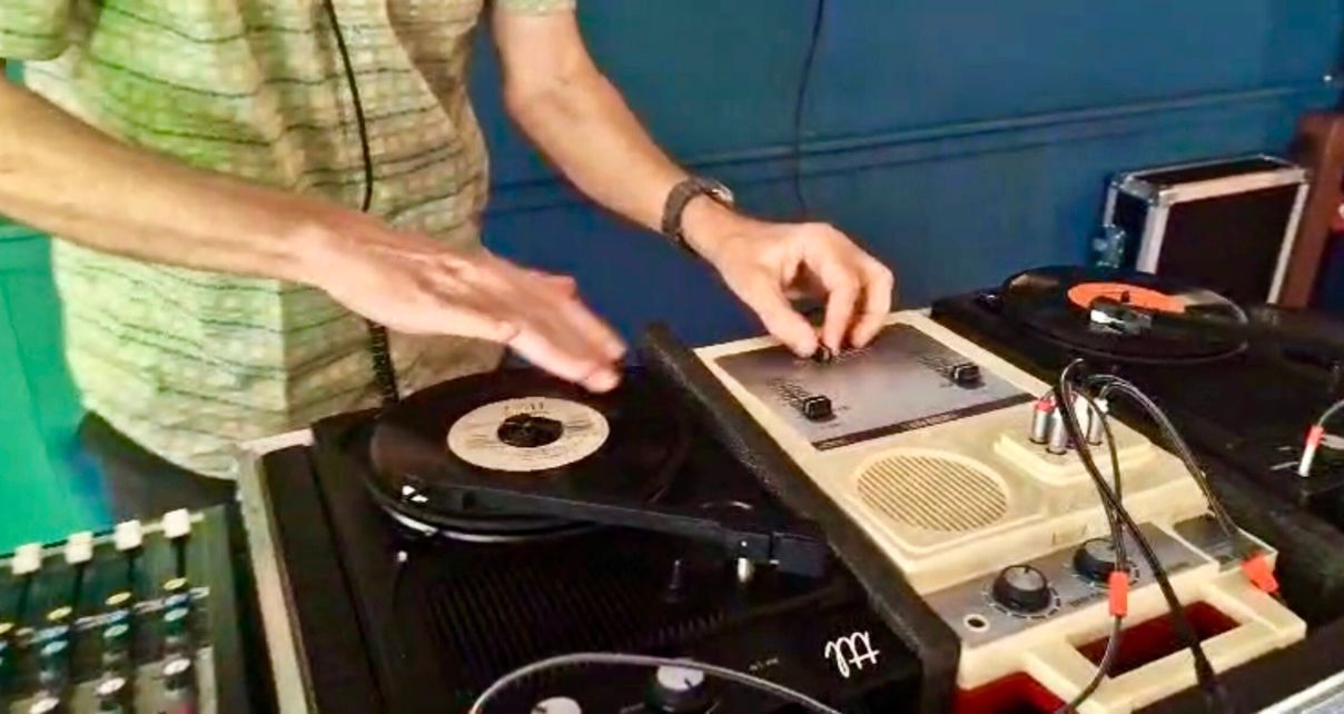 A DJ manipulates 7-inch vinyl records on turntables, focusing on one record while controlling a vintage mixer, set against a green wall.