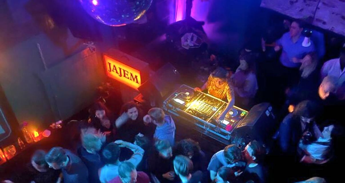 A vibrant nightclub scene featuring a DJ mixing behind a pro booth, surrounded by colourful lights and a crowd dancing energetically.