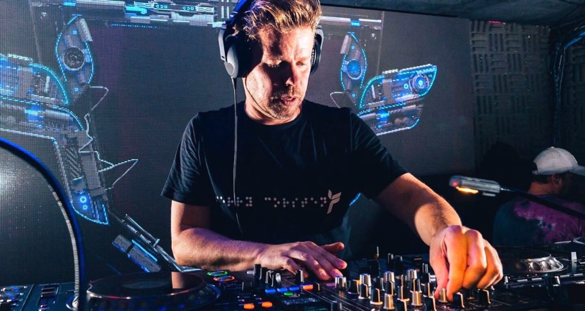 Wearing a black t-shirt and headphones, Ferry Corsten mixes music on CDJs at a club. A screen behind him projects some colourful blue electronic visuals.