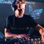 Wearing a black t-shirt and headphones, Ferry Corsten mixes music on CDJs at a club. A screen behind him projects some colourful blue electronic visuals.