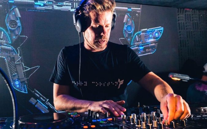 Wearing a black t-shirt and headphones, Ferry Corsten mixes music on CDJs at a club. A screen behind him projects some colourful blue electronic visuals.