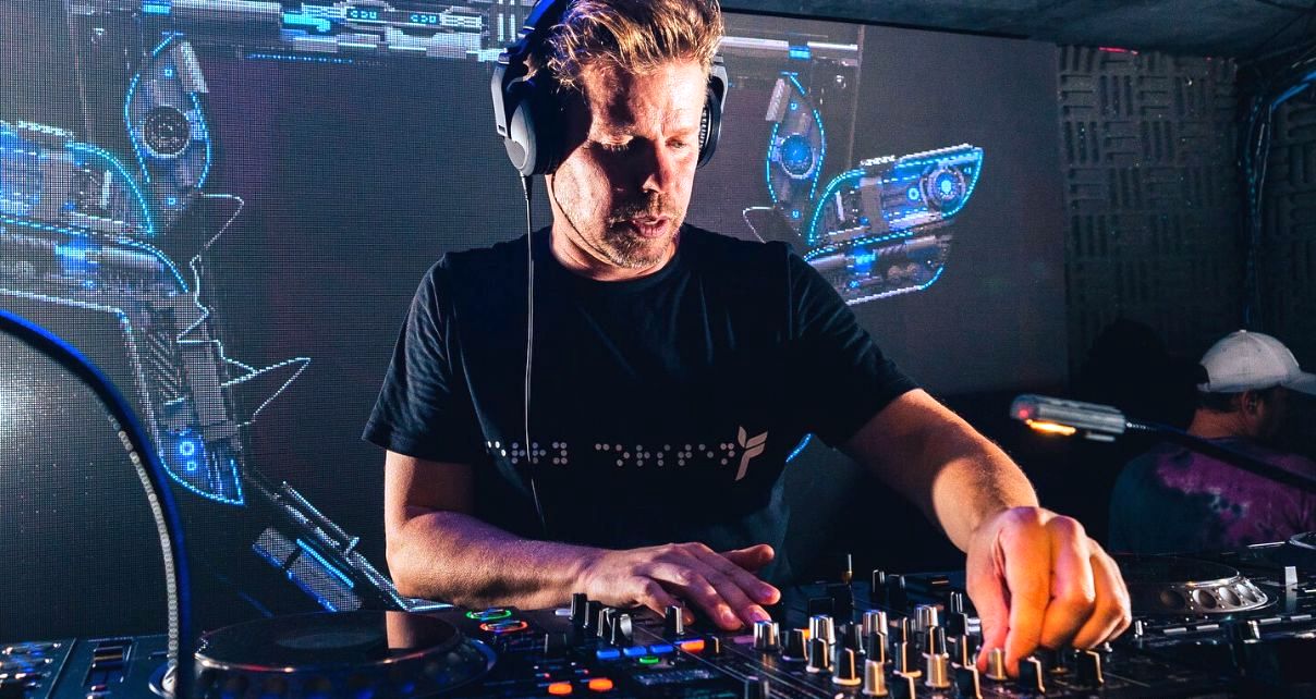 Wearing a black t-shirt and headphones, Ferry Corsten mixes music on CDJs at a club. A screen behind him projects some colourful blue electronic visuals.
