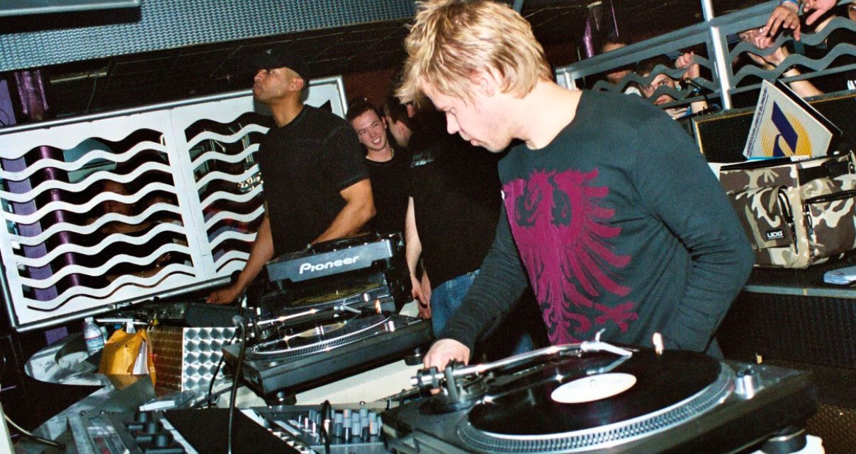 Wearing a black and red long-sleeve shirt, Ferry Corsten mixes records on turntables in a lively nightclub setting, with a crowd enjoying the music in the background.