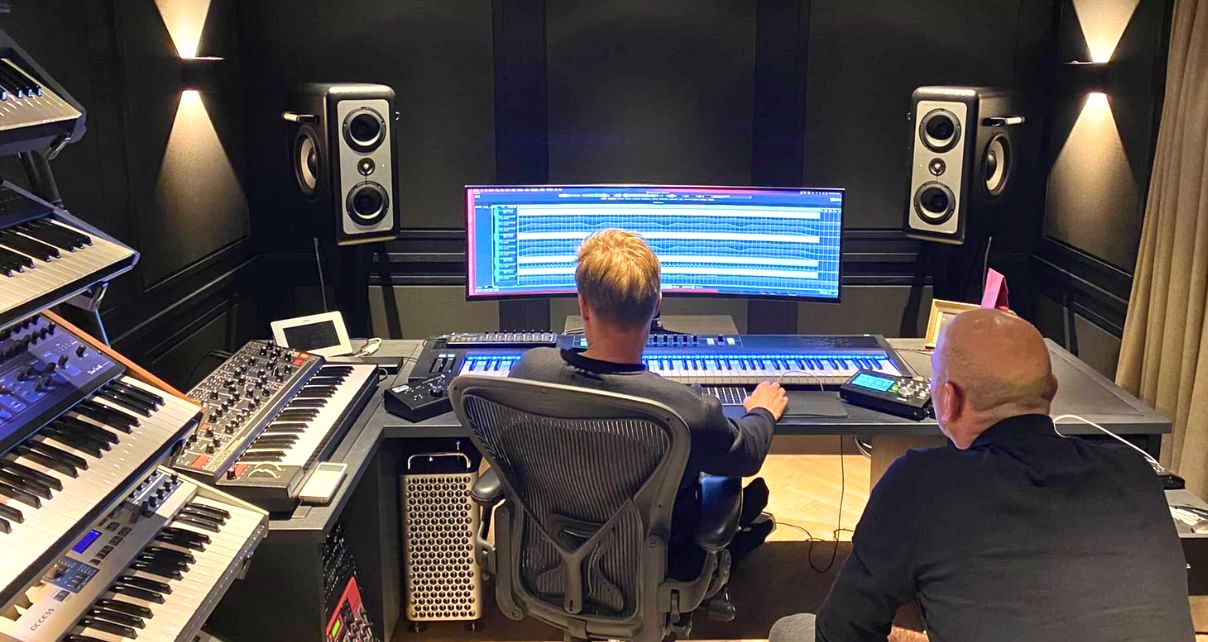 Two men sit in a dimly lit music production studio, surrounded by keyboards. One watches the large computer monitor as the other works in a DAW on a music project.