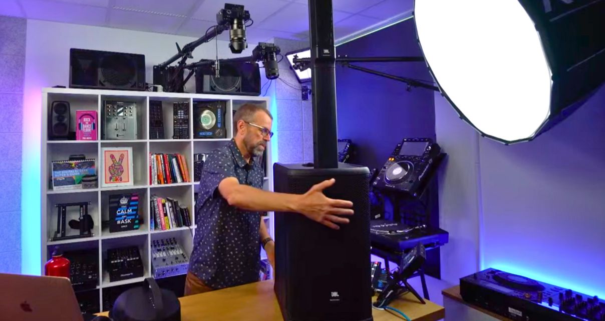 A white man with glasses, a beard, shaved hair, and a button-up shirt adjusts a tall JBL Eon One Mk2 speaker on a wooden desk. The surrounding studio is filled with audio equipment, shelves of books, and colourful lighting.