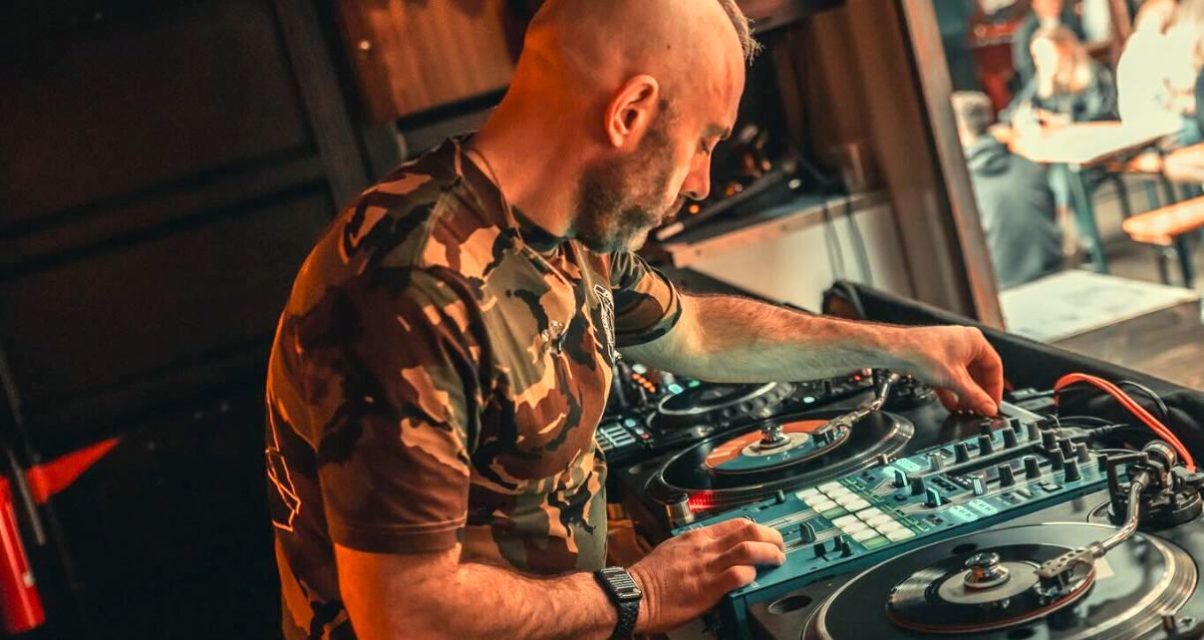 Side view of Mojaxx DJing on 7" singles. An outdoor crowd is seated and mingling off to the right.