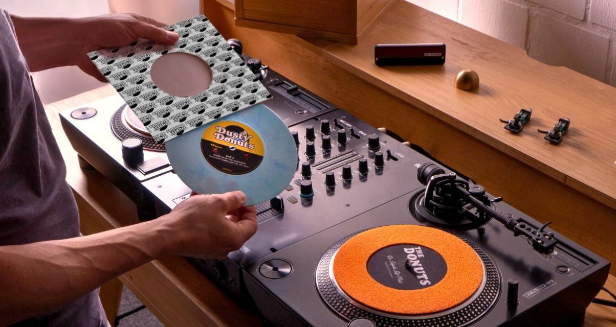 Two black Reloop RP-7 turntables and a mixer sit on a wooden desk. Two turntable needles, an adapter, and a vinyl brush sit on the shelf above. A person is taking a powder blue 7" single out of its sleeve. The label reads "Dusty Donuts".