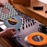 Two black Reloop RP-7 turntables and a mixer sit on a wooden desk. Two turntable needles, an adapter, and a vinyl brush sit on the shelf above. A person is taking a powder blue 7" single out of its sleeve. The label reads "Dusty Donuts".