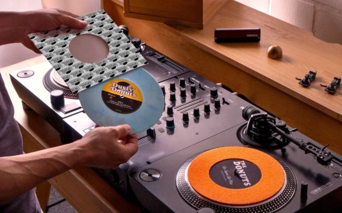 Two black Reloop RP-7 turntables and a mixer sit on a wooden desk. Two turntable needles, an adapter, and a vinyl brush sit on the shelf above. A person is taking a powder blue 7" single out of its sleeve. The label reads "Dusty Donuts".