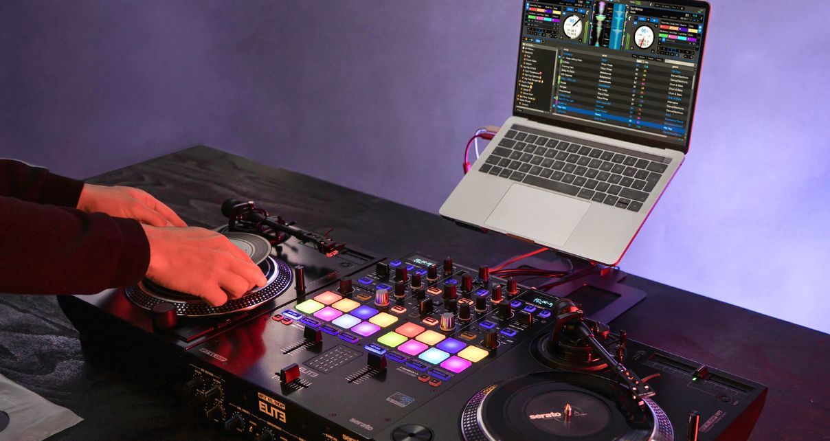 A DJ places Serato control vinyl on a Reloop RP-7 turntable. A Reloop Elite mixer and a second RP-7 with control vinyl sit next to it. Behind the decks is a silver laptop displaying Serato DJ software.